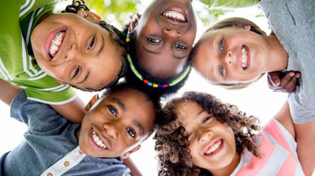 A multi-ethnic group of elementary age children are huddled together, standing in a circle with their heads in the center looking down at the camera. They are smiling and looking at the camera.